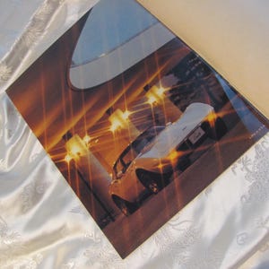 May include: A printed advertisement featuring a yellow sports car with a white top, illuminated by bright lights. The image is set against a backdrop of a building interior with a warm, golden color scheme. The advertisement is on a white satin surface.