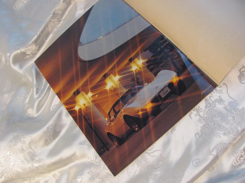May include: A printed advertisement featuring a yellow sports car with a white top, illuminated by bright lights. The image is set against a backdrop of a building interior with a warm, golden color scheme. The advertisement is on a white satin surface.