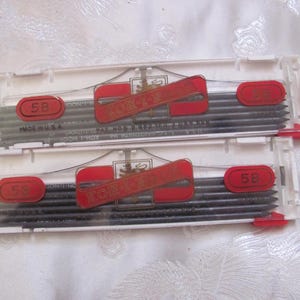 May include: Two clear plastic cases of Koh-I-Noor 5B graphite leads. Each case has a red label with the Koh-I-Noor logo and red oval stickers marked "5B". The cases are labeled "Made in U.S.A."