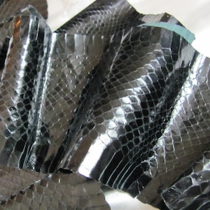 May include: Close-up of black snakeskin leather with a glossy finish. The leather has a scale pattern and is folded over itself. The material appears to be flexible and suitable for various crafting projects.