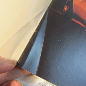 May include: A partial view of a glossy magazine page featuring a sports car. The page is being lifted to reveal the image of the car, which is orange. The background is dark.