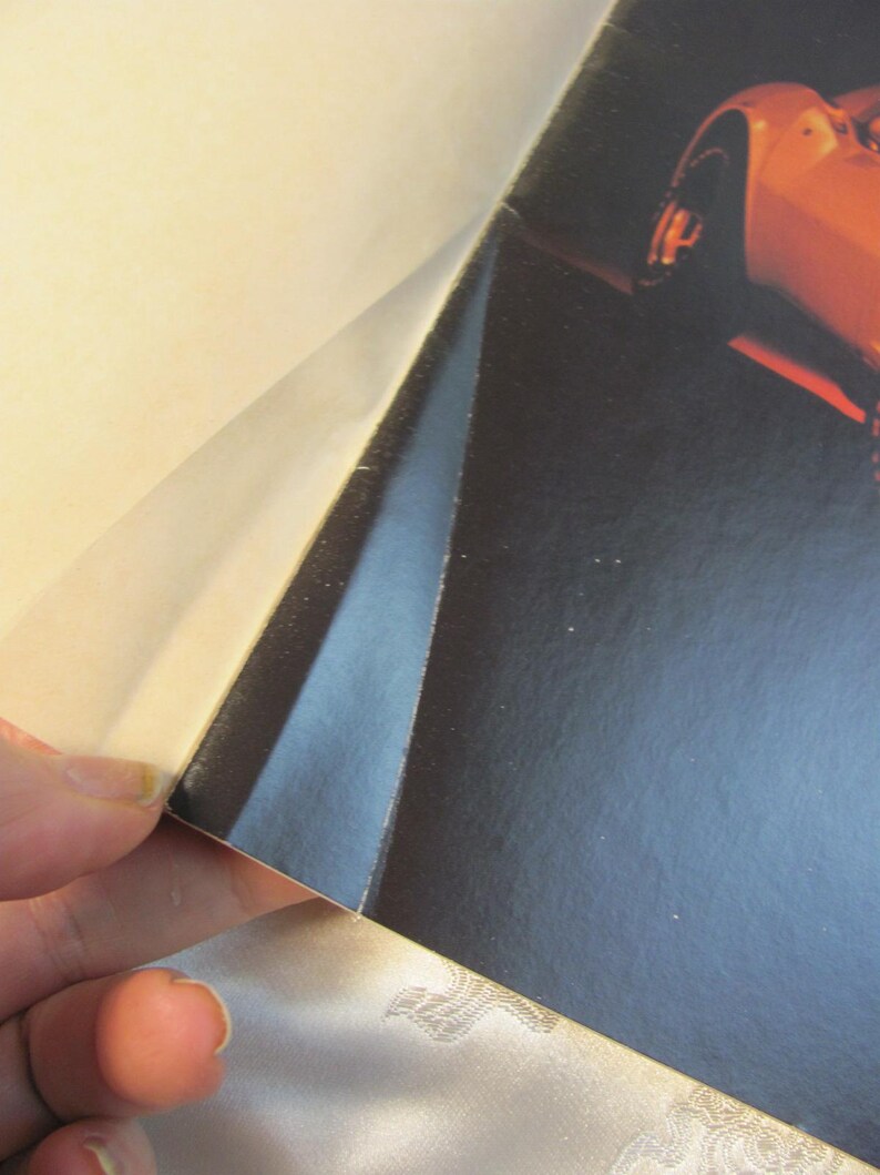 May include: A partial view of a glossy magazine page featuring a sports car. The page is being lifted to reveal the image of the car, which is orange. The background is dark.