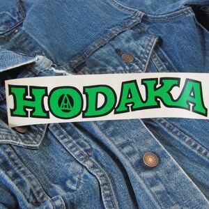 May include: A rectangular white sticker featuring the word HODAKA in bold green capital letters with a black outline. The letter O contains a small black triangular logo. The sticker is placed on a blue denim jacket with visible metal buttons.