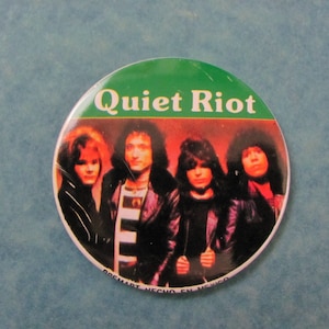 May include: A round button with a green background and the text "Quiet Riot" in white. The button features a photo of the band members, with a red, orange, and yellow background.
