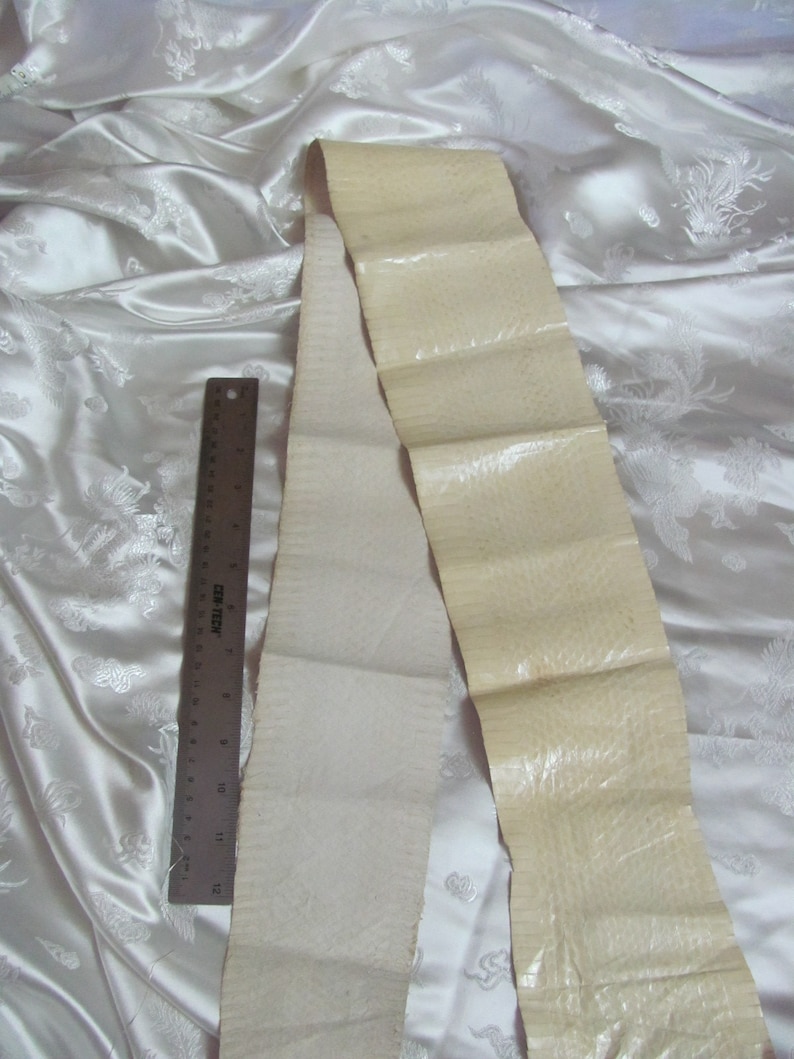 May include: Two strips of off-white leather, one with a smooth surface and the other with a textured surface, are laid out on a white fabric background. A ruler is placed next to the strips of leather.