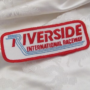 May include: A rectangular patch with the words "RIVERSIDE INTERNATIONAL RACEWAY" in red lettering. The patch has a white background with blue lines and a red border. The patch is on a white fabric.