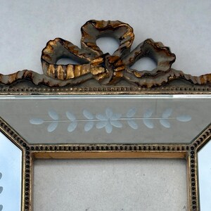 May include: A decorative picture frame with a gold-coloured trim and a bow-shaped embellishment at the top. The frame has a light-coloured background with etched floral designs. The frame is designed to hold a photograph or artwork.