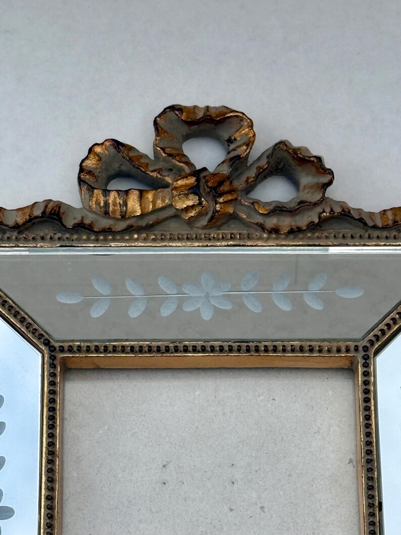 May include: A decorative picture frame with a gold-coloured trim and a bow-shaped embellishment at the top. The frame has a light-coloured background with etched floral designs. The frame is designed to hold a photograph or artwork.