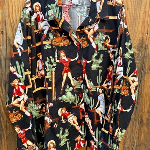May include: Black button-down shirt with a repeating pattern of cowgirls, cacti, and cowboy-themed illustrations. The shirt features a collared neckline and long sleeves. The tag reads "Sharon JS".
