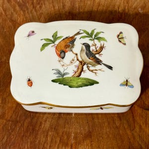 May include: A white porcelain trinket box with a scalloped edge. The lid features a detailed illustration of two birds perched on a tree branch, surrounded by butterflies and a ladybug. The box has a gold trim.