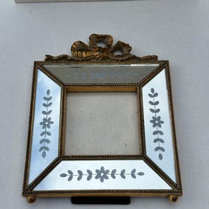 May include: A square, gold-toned picture frame with mirrored glass and etched floral designs. The frame has a decorative bow at the top and small ball feet. The frame is designed to hold a photograph.