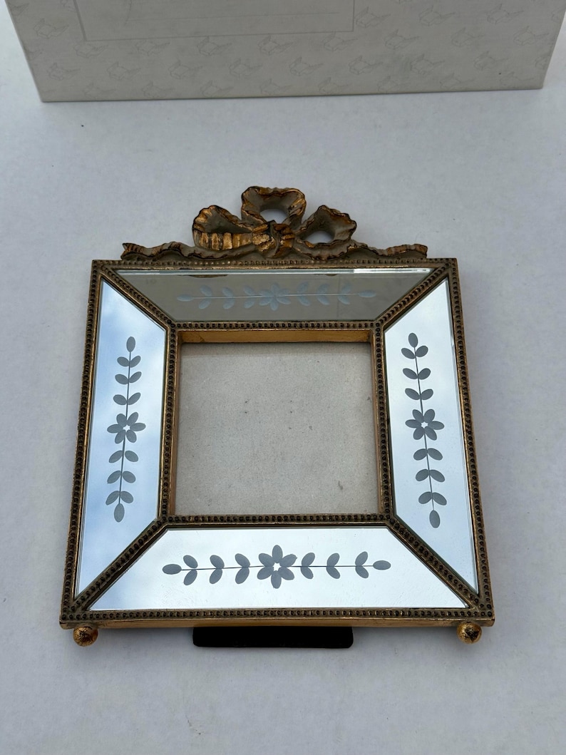 May include: A square, gold-toned picture frame with mirrored glass and etched floral designs. The frame has a decorative bow at the top and small ball feet. The frame is designed to hold a photograph.