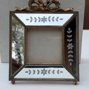 May include: A square, antique-style picture frame with a gold-coloured metal frame and a mirrored surface. The frame features etched floral designs and a decorative bow at the top. The frame is supported by small, round feet.