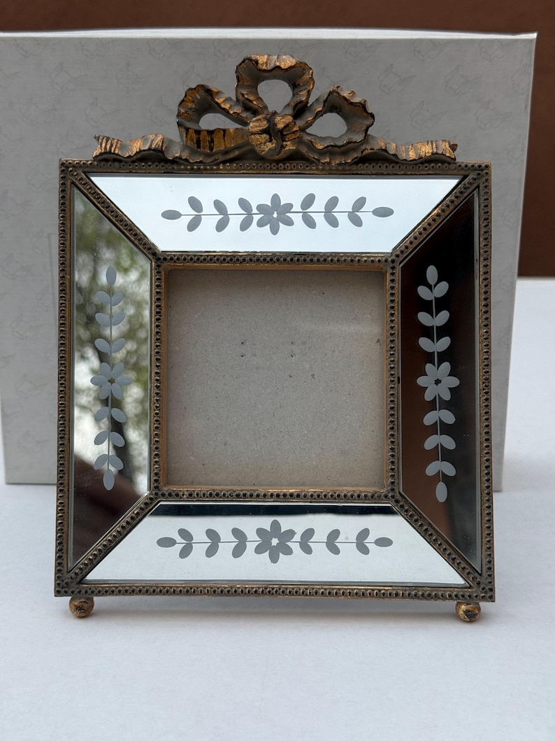 May include: A square, antique-style picture frame with a gold-coloured metal frame and a mirrored surface. The frame features etched floral designs and a decorative bow at the top. The frame is supported by small, round feet.