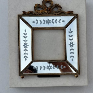 May include: A square mirror frame with a gold-coloured metal frame and a decorative bow at the top. The mirror has etched floral and leaf designs. The frame is set on a light-coloured, patterned background.