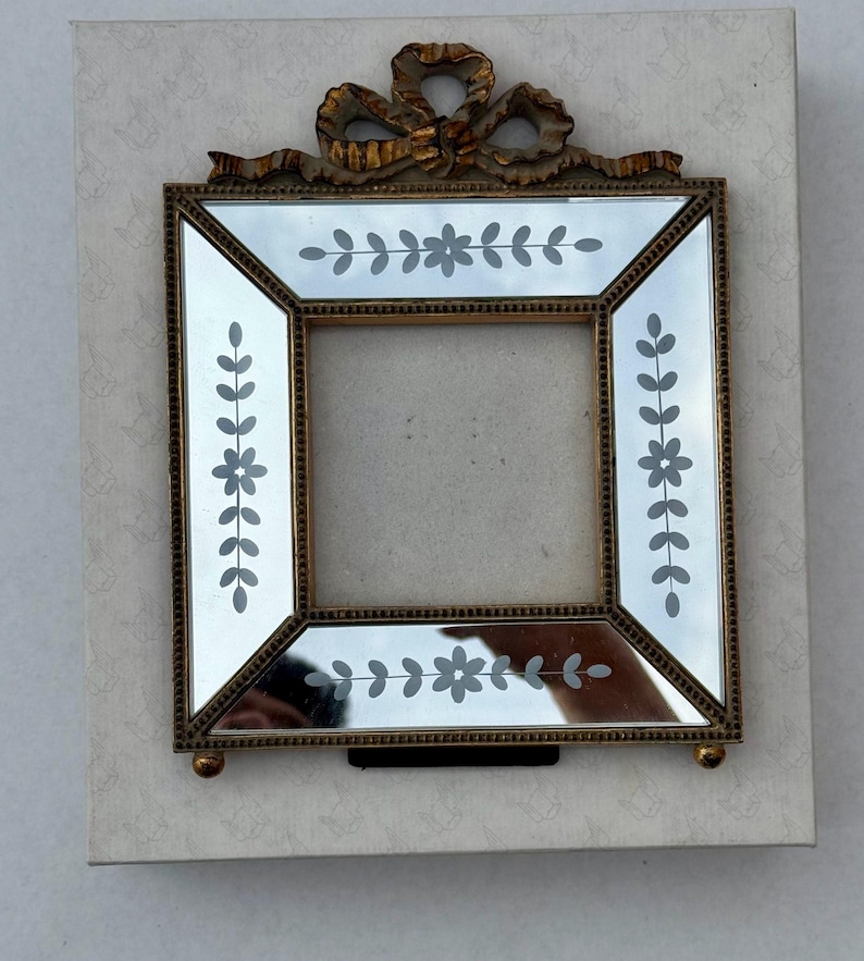 May include: A square mirror frame with a gold-coloured metal frame and a decorative bow at the top. The mirror has etched floral and leaf designs. The frame is set on a light-coloured, patterned background.