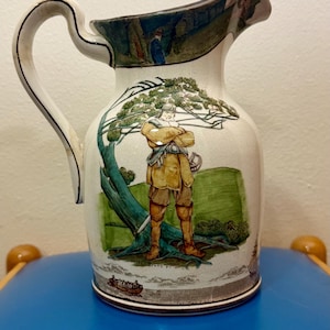 May include: Cream-colored ceramic pitcher with a curved handle and a detailed illustration. The artwork depicts a figure in period clothing standing near a tree, with a landscape scene and text at the base.
