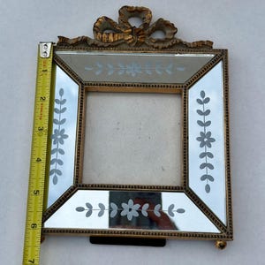 May include: A vintage-style picture frame with a gold-coloured metal frame and mirrored glass. The frame features etched floral designs and a decorative bow at the top. The frame measures approximately 15 cm in height.