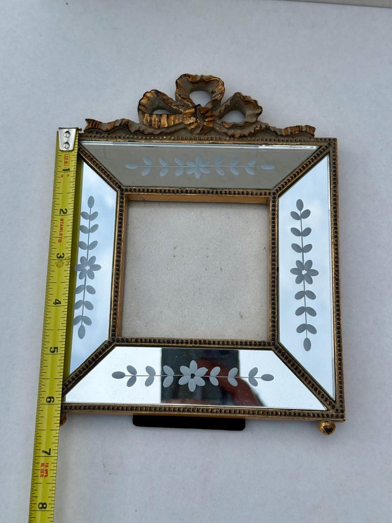 May include: A vintage-style picture frame with a gold-coloured metal frame and mirrored glass. The frame features etched floral designs and a decorative bow at the top. The frame measures approximately 15 cm in height.