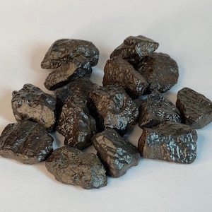 Könnte beinhalten: Eine Ansammlung dunkelbrauner, unregelmäßig geformter Steine mit strukturierter Oberfläche. Die Steine sind zusammengefasst und füllen den Rahmen vor einem schlichten weißen Hintergrund. Die Steine scheinen unterschiedliche Größen und Formen zu haben.