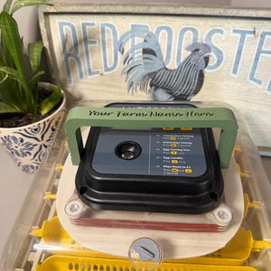 May include: An egg incubator featuring a clear plastic lid, a black control panel with instructional text, and a sage green handle with the text "Your Farm Name Here" printed on it. The interior contains yellow plastic egg turning trays.