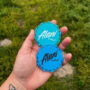 Puede incluir: Dos posavasos redondos, verde azulado y azul, con borde festoneado. Cada posavasos presenta el nombre "Alani" en una fuente de escritura blanca. Los posavasos se sostienen en una mano sobre un fondo de hierba verde.