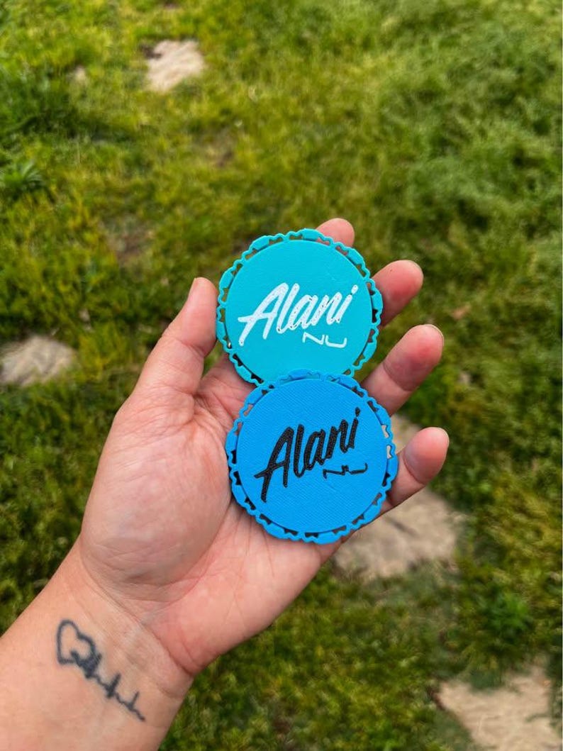 Puede incluir: Dos posavasos redondos, verde azulado y azul, con borde festoneado. Cada posavasos presenta el nombre "Alani" en una fuente de escritura blanca. Los posavasos se sostienen en una mano sobre un fondo de hierba verde.