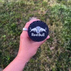 Puede incluir: Un posavasos redondo negro con el logotipo de Red Bull en blanco. El posavasos est&aacute; sujeto por un anillo plateado. El fondo es hierba verde borrosa.