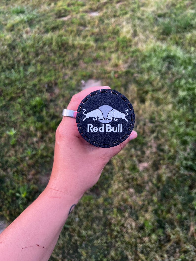 Puede incluir: Un posavasos redondo negro con el logotipo de Red Bull en blanco. El posavasos est&aacute; sujeto por un anillo plateado. El fondo es hierba verde borrosa.