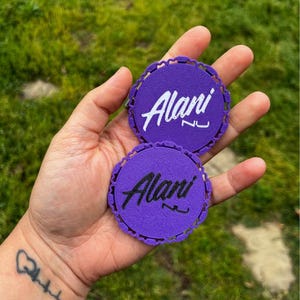 Puede incluir: Dos parches circulares morados con bordes festoneados, cada uno con el nombre "Alani" en escritura blanca. Los parches se sostienen en una mano, con hierba verde en el fondo.