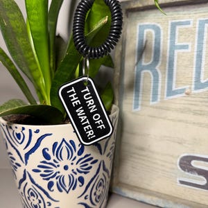 May include: A black plastic coil wristband attached to a rectangular black tag with white border and white text that reads TURN OFF THE WATER! The tag is connected to the coil by a small silver metal split ring, creating a functional keychain.