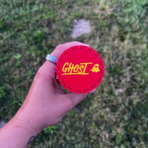 Puede incluir: Un objeto circular rojo con la palabra "GHOST" en amarillo y un gr&aacute;fico de fantasma amarillo. El objeto tiene un borde cosido y se sostiene sobre un fondo de hierba verde.