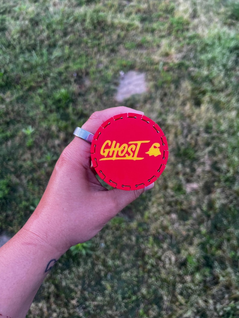 Puede incluir: Un objeto circular rojo con la palabra "GHOST" en amarillo y un gr&aacute;fico de fantasma amarillo. El objeto tiene un borde cosido y se sostiene sobre un fondo de hierba verde.