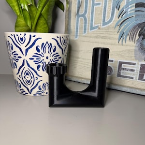 May include: A black U-shaped plumbing pipe fitting with a threaded collar on one side. The component features a smooth, solid finish and a distinct curved base, designed for connecting drainage sections in a standard plumbing configuration.