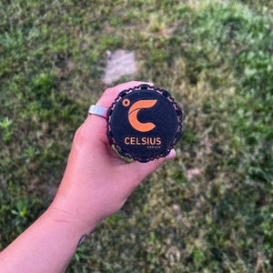 Puede incluir: Una tapa negra y naranja de una lata de bebida energ&eacute;tica Celsius se sostiene en una mano. La tapa tiene el logotipo y el texto de Celsius. El fondo es hierba verde.