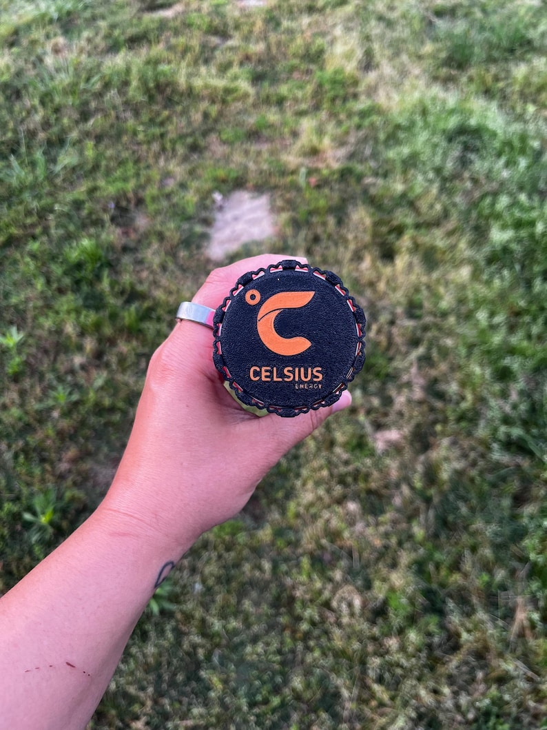 Puede incluir: Una tapa negra y naranja de una lata de bebida energ&eacute;tica Celsius se sostiene en una mano. La tapa tiene el logotipo y el texto de Celsius. El fondo es hierba verde.