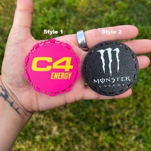 Puede incluir: Dos posavasos redondos. Uno es de color rosa brillante con el texto amarillo "C4 ENERGY". El otro es negro con el logotipo blanco "Monster Energy". Ambos tienen un borde decorativo.