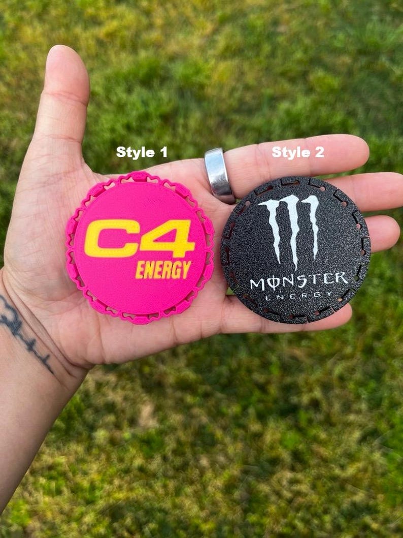 Puede incluir: Dos posavasos redondos. Uno es de color rosa brillante con el texto amarillo "C4 ENERGY". El otro es negro con el logotipo blanco "Monster Energy". Ambos tienen un borde decorativo.