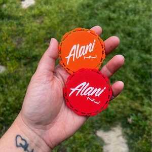 Puede incluir: Dos posavasos redondos, naranja y rojo, sostenidos en una mano. Cada uno tiene un borde festoneado y el nombre "Alani" en escritura blanca. El fondo es hierba verde borrosa.