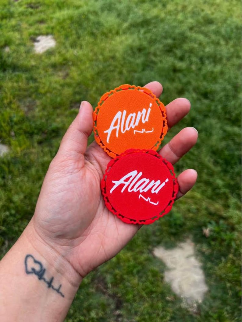 Puede incluir: Dos posavasos redondos, naranja y rojo, sostenidos en una mano. Cada uno tiene un borde festoneado y el nombre "Alani" en escritura blanca. El fondo es hierba verde borrosa.