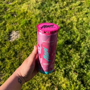 Puede incluir: Una lata rosa de bebida energ&eacute;tica Alani Nu con tapa rosa y base turquesa. La lata presenta el logotipo de Alani Nu y las palabras "Pink Slush" y "Energy". La lata se sostiene en una mano sobre un fondo de hierba verde.