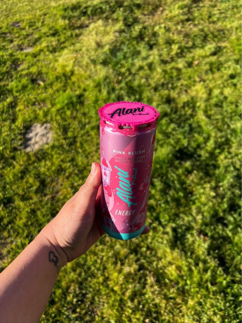 Puede incluir: Una lata rosa de bebida energ&eacute;tica Alani Nu con tapa rosa y base turquesa. La lata presenta el logotipo de Alani Nu y las palabras "Pink Slush" y "Energy". La lata se sostiene en una mano sobre un fondo de hierba verde.