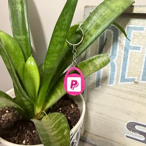 May include: A bright pink glittery keychain featuring an oval shape with a cutout handle at the top. The center of the keychain displays a white square with a bold pink letter P printed on it. It is attached to a silver metal split ring.