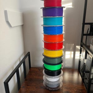 Puede incluir: Una pila alta de bobinas de filamento de impresora 3D de colores. Las bobinas son de varios colores, incluyendo rojo, morado, turquesa, azul, naranja, amarillo, verde, negro, blanco y gris. Las bobinas est&aacute;n apiladas sobre una superficie de madera marr&oacute;n oscuro.