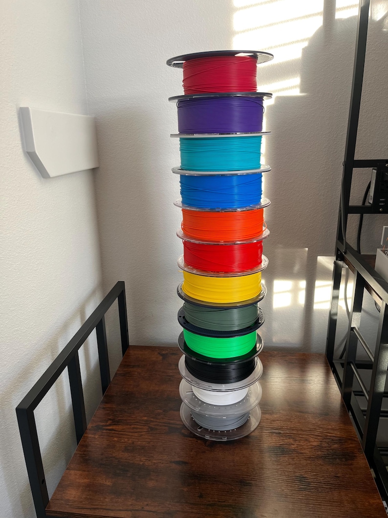 Puede incluir: Una pila alta de bobinas de filamento de impresora 3D de colores. Las bobinas son de varios colores, incluyendo rojo, morado, turquesa, azul, naranja, amarillo, verde, negro, blanco y gris. Las bobinas est&aacute;n apiladas sobre una superficie de madera marr&oacute;n oscuro.