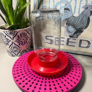May include: A clear glass jar inverted into a bright red plastic base, which sits atop a circular, vibrant pink platform featuring a repeating pattern of small, dark, teardrop-shaped cutouts arranged in a radial design.