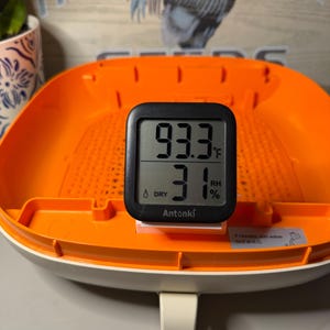 May include: A black digital Antonki thermometer and hygrometer displaying 93.3 degrees Fahrenheit and 31 percent relative humidity, positioned inside a bright orange plastic tray with a perforated base and a small instructional sticker.