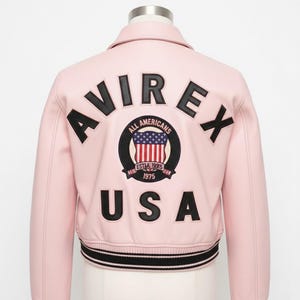 Könnte beinhalten: Hellrosa Lederjacke mit dem Wort "AVIREX" und "USA" auf dem Rücken. Ein kreisförmiger Aufnäher mit der amerikanischen Flagge und den Worten "ALL AMERICANS EST. 1975" ist zentriert. Die Jacke hat einen schwarz-weiß gestreiften Besatz.
