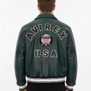 Könnte beinhalten: Dunkelgrüne Lederjacke mit dem Wort "AVIREX" auf dem Rücken, über einem runden Aufnäher mit der amerikanischen Flagge und den Worten "ALL AMERICANS 1975". Das Wort "USA" steht darunter. Die Jacke hat einen weiß-schwarz gestreiften Saum.