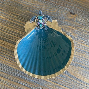 Könnte beinhalten: Eine türkisblaue, muschelförmige Schmuckschale mit goldfarbenem Rand. Eine mit Juwelen besetzte Schildkrötenfigur ist an der Schale befestigt. Die Schildkröte hat blaue und irisierende Edelsteine. Die Schale steht auf einer Holzoberfläche.